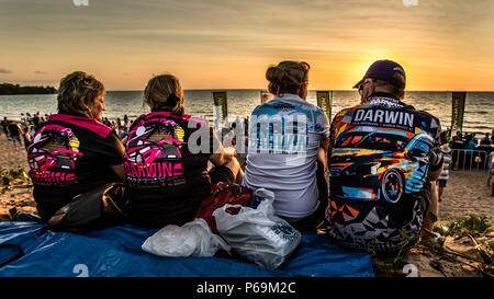 Four visitors wearing jerseys from the Darwin Supercars race weekend sit with their backs to the camera at Mindil Beach in Darwin, Northern Territory, Australia, watching the sunset over the sea. Stock Photo