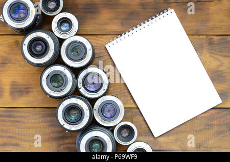 Several photographic lenses and white notebook lie on a brown wooden background. Space for text . Stock Photo