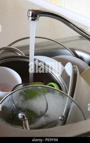 Dirty dishes and unwashed kitchen appliances lie in foam water under a tap from a kitchen faucet . Stock Photo