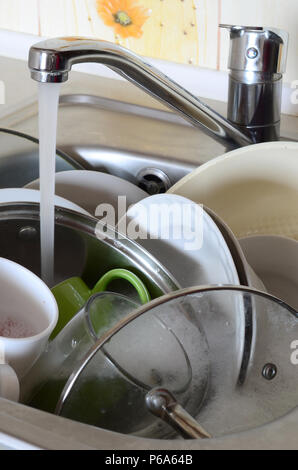 Dirty dishes and unwashed kitchen appliances lie in foam water under a tap from a kitchen faucet . Stock Photo