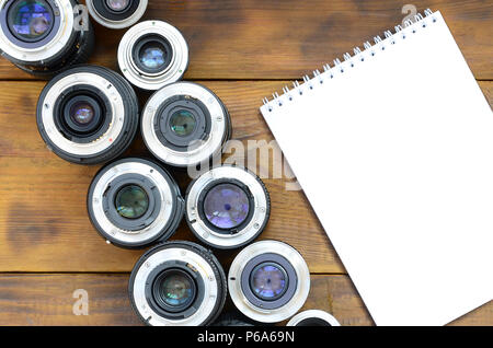 Several photographic lenses and white notebook lie on a brown wooden background. Space for text . Stock Photo