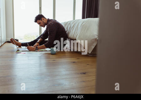 Man working on laptop computer sitting on the floor. Freelancer sitting on floor beside a bed and making notes while working on laptop at home with a Stock Photo