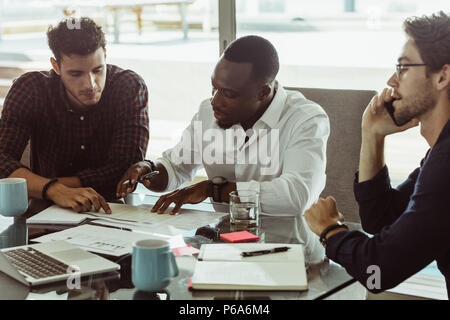 Businessmen discussing work sitting at conference table in office. Two men discussing work while another man is talking on mobile phone. Stock Photo