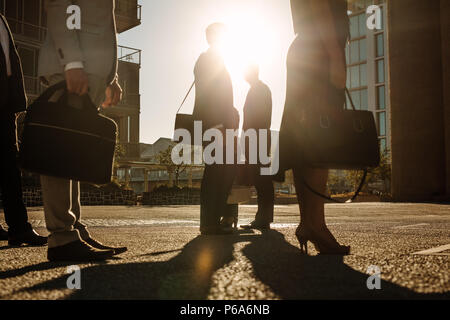 Men and woman in formal clothes commuting to office in the morning carrying office bags. Office going people standing on a busy street with sun flare  Stock Photo