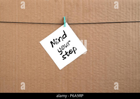 Mind your step. Note is written on a white sticker that hangs with a clothespin on a rope on a background of brown cardboard . Stock Photo