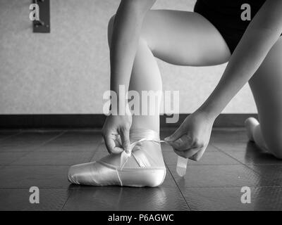 Black and white version of Recreational young female ballet dancer ballerina, in the studio putting on her pointe shoes, tying up her ribbons Stock Photo