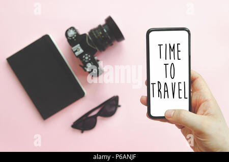 Time to Travel text on screen,  hand holding stylish black phone , sunglasses, photo camera and notebook on pink background, flat lay. instagram blogg Stock Photo