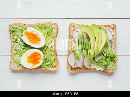 Two avocado toasts with soft boiled eggs,mashed avocado,sliced avocado ,radish, snow pea sprouts and goat cheese on white wooden background Stock Photo