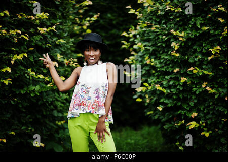 Amazing african american model woman in green pants and black hat posed with different emotions at park, shows a two fingers. Stock Photo
