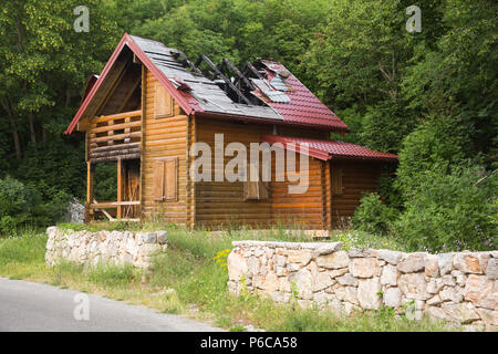 Abandoned badly fire damaged wooden house with gaping hole in roof on mountainside at Njeguši above Kotor, Montenegro with green trees in background. Stock Photo