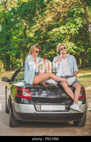 Beautiful fashionable couple sitting at the back of convertible car and using their smart phones. Stock Photo