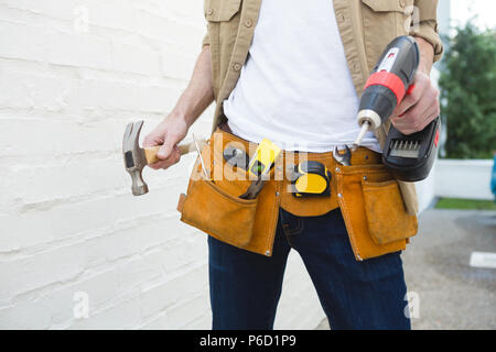 Male carpenter with tool belt holding hammer and drill machine Stock Photo