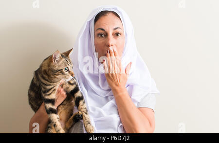 Arabian woman wearing white hijab holding a cat cover mouth with hand shocked with shame for mistake, expression of fear, scared in silence, secret co Stock Photo