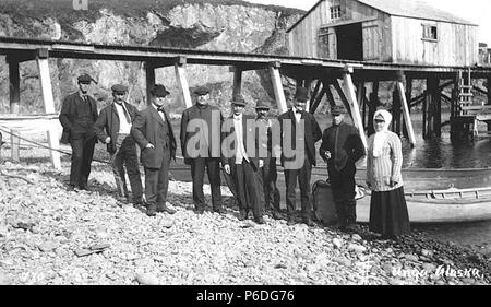 . English: Group of people standing on shore near boat dock, Squaw Harbor, Unga Island, ca. 1912 . English: Caption on image: Unga, Alaska PH Coll 247.221 Unga lies on the southeast coast of Unga Island, in the Shumagin Islands of the Aleutian Chain. It was an Aleut village first reported as 'Delarov' in 1833, with a population of 116, and then as 'Ougnagok' in 1836. The 'Ounga' post office was established in 1888, and changed its name to Unga in 1894. The post office closed in 1958. Unga is no longer occupied year-round. Subjects (LCTGM): Squaw Harbor (Alaska); Unga Island(Alaska); Piers & wh Stock Photo