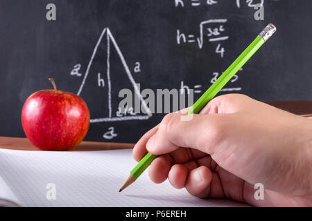 Male student hand writes in notebook with green wooden pen. Red apple and blackboard is blurred in background. Classroom and education concept Stock Photo