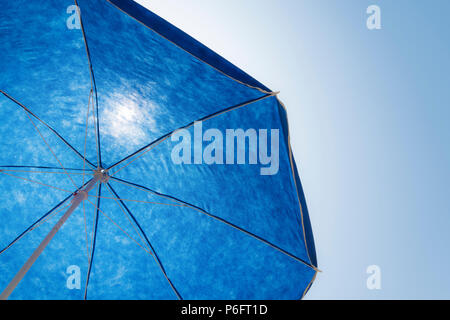 Under a bright sunny sky, a blue beach parasol is shown in contre-jour, emphasizing its silhouette against the clear, blue backdrop Stock Photo