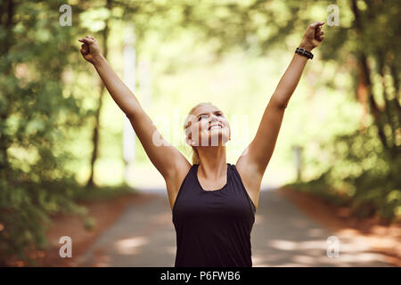 Fit young woman in sportwear smiling while standing on a forest path with her arms raised in achievement after a run Stock Photo