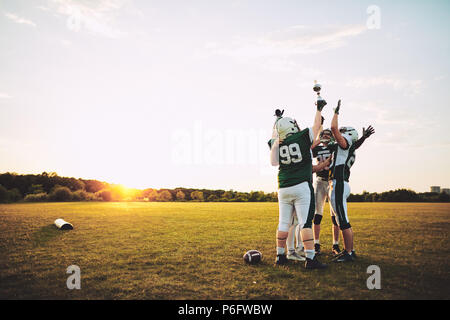 Ecstatic group of American football players standing in a huddle and raising a championship trophy in celebration Stock Photo