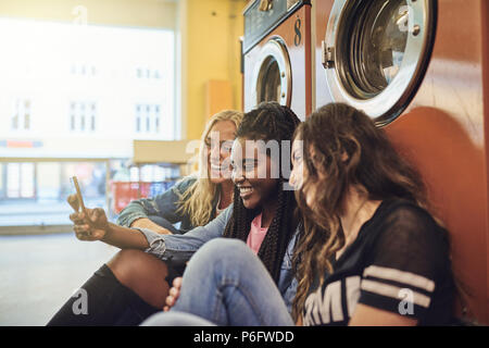 Diverse young female friends laughing and sitting together on a laundromat floor using a cellphone Stock Photo