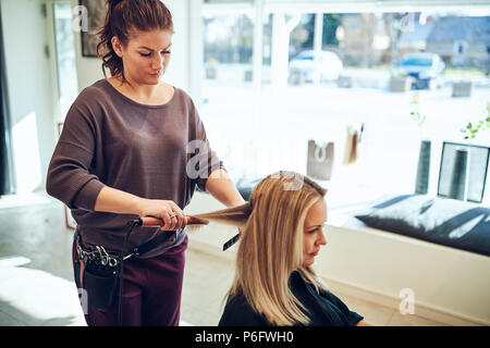 Young blonde woman sitting in a salon chair getting her long blonde hair straightened by her hairstylist Stock Photo