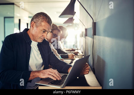 Focused mature designer sitting at a table with coworkers in a modern office working online with a laptop Stock Photo