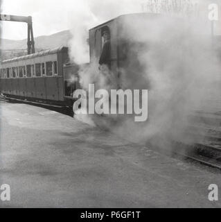 1950s, steam engulfs the driver in his cabin as he enjoy's a cigarette as the small tourist locomotive waits on the platform at the Snowdon mountain railway, Wales, UK. Stock Photo