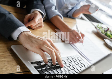 Man and woman having business lunch at restaurant sitting at table businessman pushing button on laptop while businesswoman taking notes on document c Stock Photo
