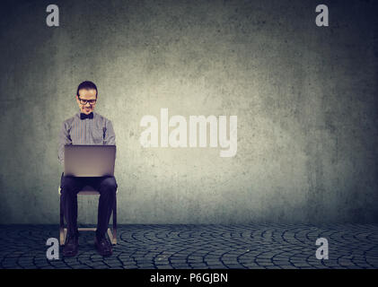 Smiling formal man in glasses sitting on chair and using laptop against gray background working in Internet Stock Photo