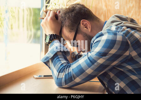 Young depressed man sitting at table with mobile phone in cafe looking down being unhappy with breakup. Stock Photo