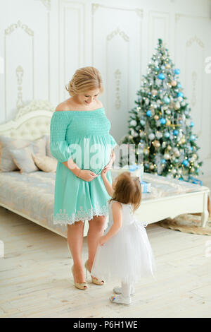 Pregnant caucasian woman wearing blue dress hugging belly and standing with little daughter near Christmas tree in bedroom. Stock Photo