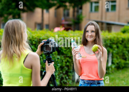Two teenage girls. Summer in nature. In her hands holds bottle of water and an apple. The concept of healthy eating. Young bloggers record video for t Stock Photo