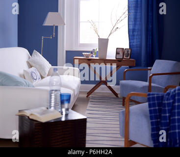 White sofa and pale blue upholstered chairs in a blue living room with a tall white vase on a wooden table in front of the window Stock Photo