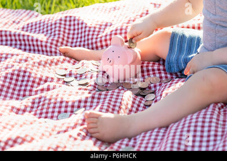 Baby Boy Sitting on Picnic Blanket PUtting Coins in Piggy Bank Stock Photo