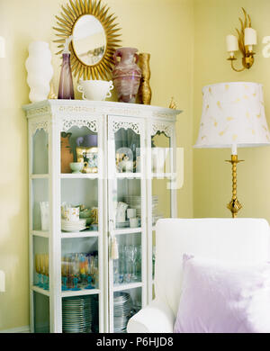Sunburst mirror and glass vases on white painted glass-front cupboard in l yellow living room Stock Photo