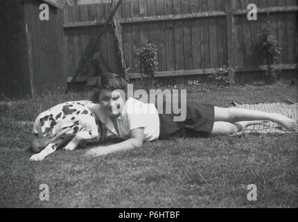 1950s, young schoolgirl lying down outside in a garden on a rug with her pet dalmatian dog. A unqiue spotted breed, Dalamations are good family dogs, being friendly, intelligent and playful. Stock Photo