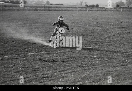 1970s, young boy wearing helmet and jacket riding outside on a flat  grass field on a scrambling motorcycle, England, UK. Stock Photo