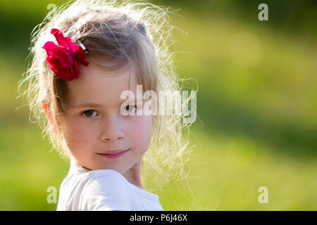 Portrait of beautiful small preschool blond girl with nice gray eyes and red rose in hair smiling dreamily in camera on blurred bright green backgroun Stock Photo