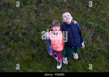 View from above of two cute children in casual clothes, boy and girl standing on green grass, hugging each other over shoulders, looking up and happil Stock Photo