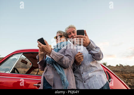 Happy elderly senior couple use mobile phones outdoor near a red beautiful retro car. leisure activity and travel together forever concept. modern use Stock Photo