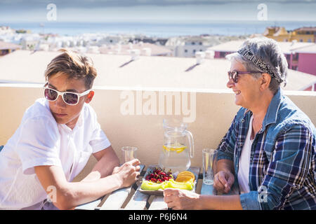 Happy and smiled couple teenager boy and grandmother female. beautiful people outdoor having breakfast in the morning on a terrace with ocean and buil Stock Photo