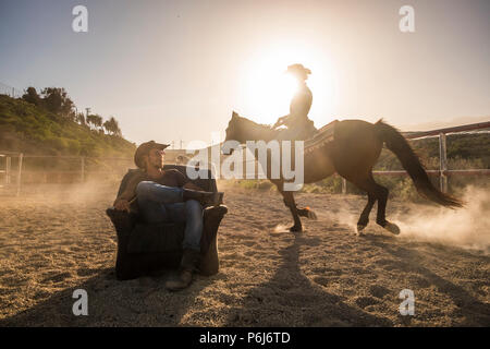 riders with horses in the golden sunset light. a man sitting on an old seat and a woman ride around him making dust. scenery image with windmills in b Stock Photo