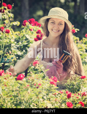 Smiling young girl in hat gardening with roses in yard Stock Photo