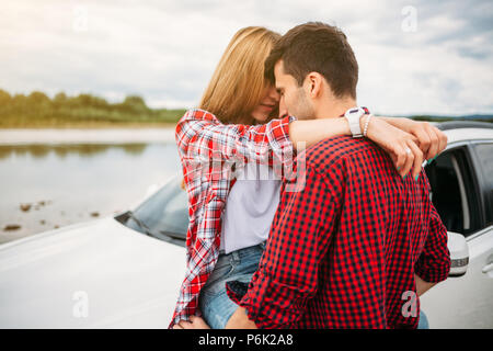 Romantic young couple sitting on the hood of their car. Beautiful young woman embraces man for neck. close up by the river Stock Photo