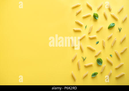Penne Pasta on yellow background, top view, flat lay. Pattern uncooked penne pasta with herbs basil and rosemary. Stock Photo