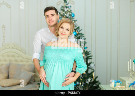 Pregnant caucasian woman wearing blue dress standing with husband near Christmas tree. Stock Photo