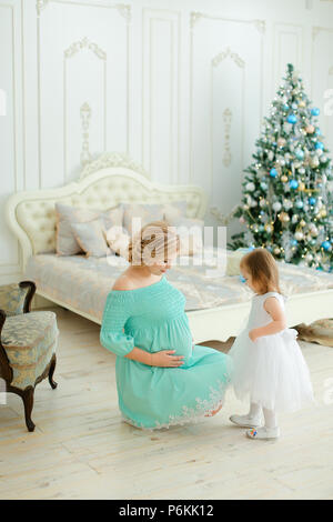 Pregnant blonde woman wearing blue dress hugging belly and sitting with little daughter near Christmas tree in bedroom. Stock Photo