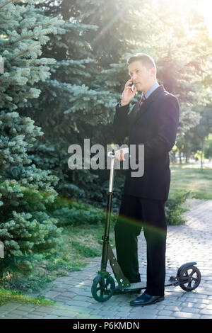 Portrait of a businessman talking on the phone while standing at the kick scooter in the park Stock Photo