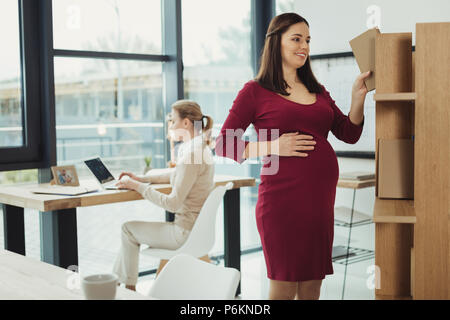 Pregnant woman smiling while taking a book from the shelf Stock Photo