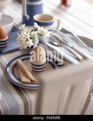 Close-up of boiled eggs in blue striped egg cups on matching plates on a striped tablecloth with a vase of scented jonquils