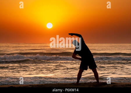 Silhouette of young man doing fitness exercise on the beach at sunrise or sunset Stock Photo